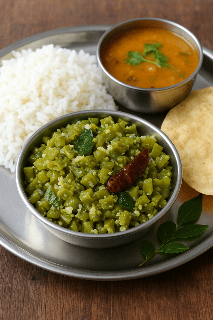 A serving of Green Bean Poriyal on a stainless steel plate, accompanied by a bowl of sambar, a mound of steamed white rice, a crispy papad, and a few curry leaves.