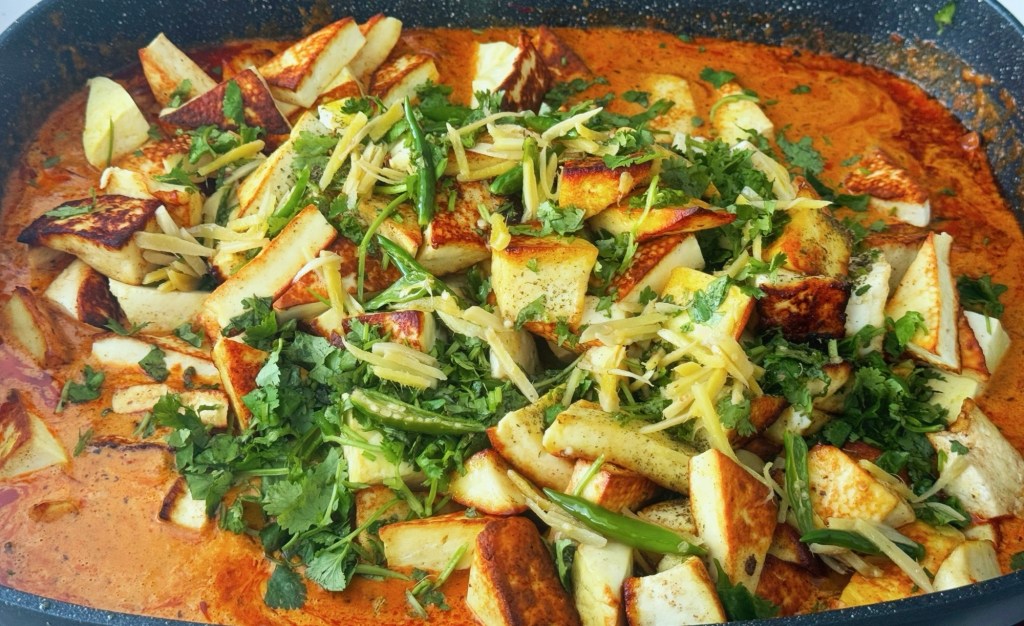 A dish of Paneer Butter Masala featuring golden-brown cubes of paneer simmered in a rich, creamy tomato sauce, topped with fresh coriander and sliced ginger.