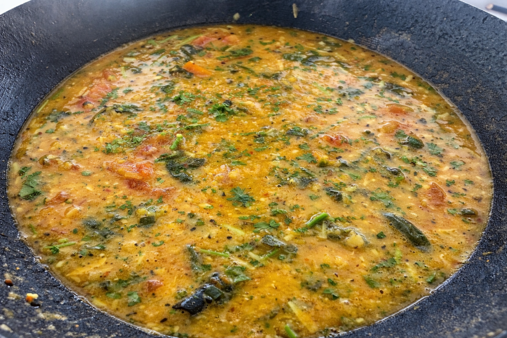 A close-up view of a pot filled with a thick, flavorful curry, featuring green herbs and tomatoes, simmering on a stove.