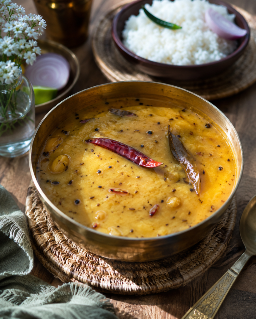 A bowl of yellow dal garnished with red chilies and spices, accompanied by a side of plain rice and sliced onions on a wooden table.